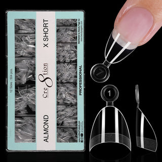 Open mint green box of clear Cre8tion Almond Extra Short tips on black, one tip fitted to a fingertip.