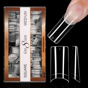 Kraft box of clear Cre8tion square tips on black, medium length, with one tip worn on a fingertip.