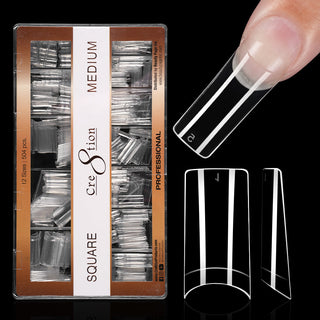 Kraft box of clear Cre8tion square tips on black, medium length, with one tip worn on a fingertip.