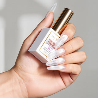 Hand with long pearly nails holding a gold capped Cre8tion Sheer Silver cat eye gel bottle by its box.